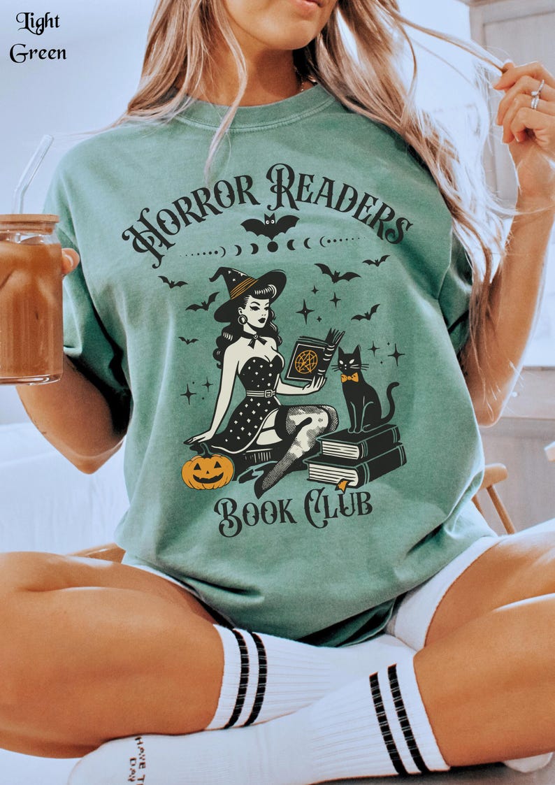 May include: A light green t-shirt with a black graphic of a witch holding a book with a black cat sitting on a stack of books. The text on the t-shirt reads 'Horror Readers Book Club'.