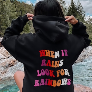 May include: A black hooded sweatshirt featuring the text "WHEN IT RAINS LOOK FOR RAINBOWS" printed on the back in a wavy, multi-colored font. The letters are colored in shades of red, orange, pink, and white, creating a vibrant, layered appearance.