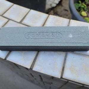 Puede incluir: Una piedra de afilar rectangular gris con el texto en relieve "CAMAHAD" en la superficie superior. La piedra tiene una base negra y est&aacute; dise&ntilde;ada para afilar herramientas. La imagen muestra la piedra sobre una superficie embaldosada.