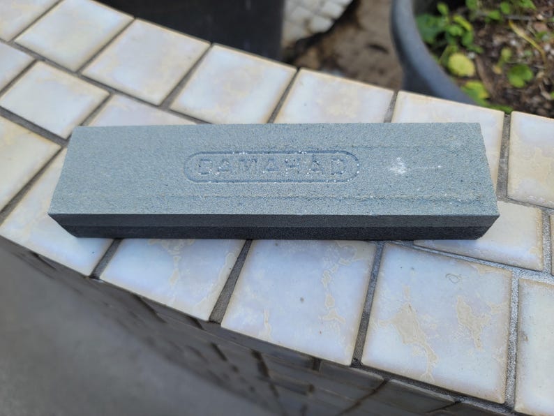 Puede incluir: Una piedra de afilar rectangular gris con el texto en relieve "CAMAHAD" en la superficie superior. La piedra tiene una base negra y est&aacute; dise&ntilde;ada para afilar herramientas. La imagen muestra la piedra sobre una superficie embaldosada.