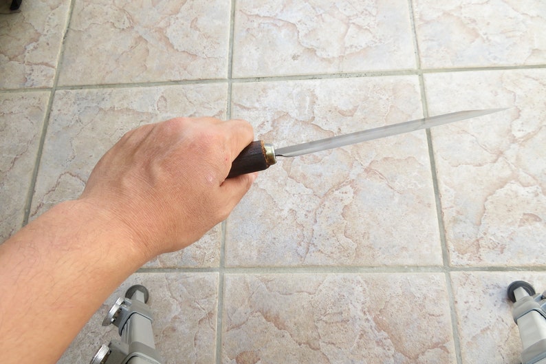 May include: A person holding a long, thin, metal file with a wooden handle. The file is approximately 30 cm long and has a slightly curved blade.