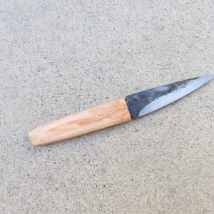 May include: A small fixed-blade knife featuring a pointed, dark-toned, forged metal blade with a visible textured finish. The handle is crafted from light-colored, smooth wood with a natural grain pattern, showing a tapered, ergonomic shape.