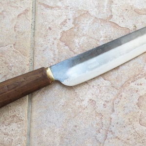 Peut inclure: Un couteau long et fin, en argent, avec un manche en bois. Le couteau a une lame l&eacute;g&egrave;rement incurv&eacute;e et un accent en laiton &agrave; la base de la lame.
