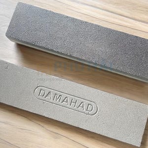 Puede incluir: Dos piedras de afilar grises. La piedra superior tiene una superficie rugosa y texturizada. La piedra inferior tiene la palabra "DAMAHAD" en relieve. Ambas piedras son rectangulares y descansan sobre una superficie de madera.
