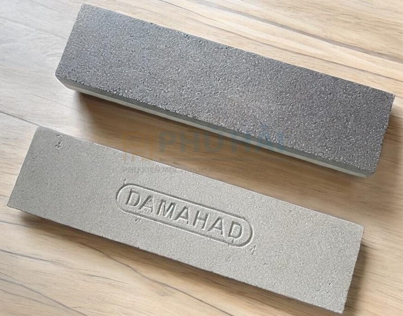 Puede incluir: Dos piedras de afilar grises. La piedra superior tiene una superficie rugosa y texturizada. La piedra inferior tiene la palabra "DAMAHAD" en relieve. Ambas piedras son rectangulares y descansan sobre una superficie de madera.