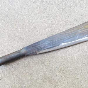 May include: A dark gray metal tool featuring a long, flat, tapered blade with a smooth, polished surface and a cylindrical handle attachment at one end. The metal shows subtle variations in tone and texture across its elongated, wedge-like shape.