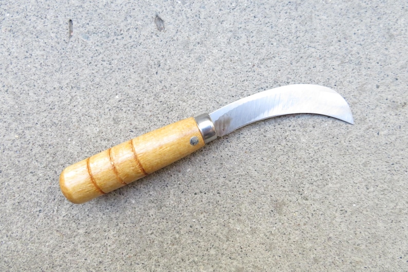 May include: A curved blade knife with a wooden handle. The blade is made of shiny metal and has a curved shape. The handle is light brown with three carved rings. The knife is designed for gardening or other cutting tasks.