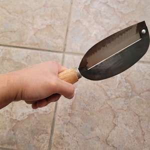 May include: A hand holds a wooden handled metal vegetable peeler with a silver blade. The peeler is on a tiled surface.