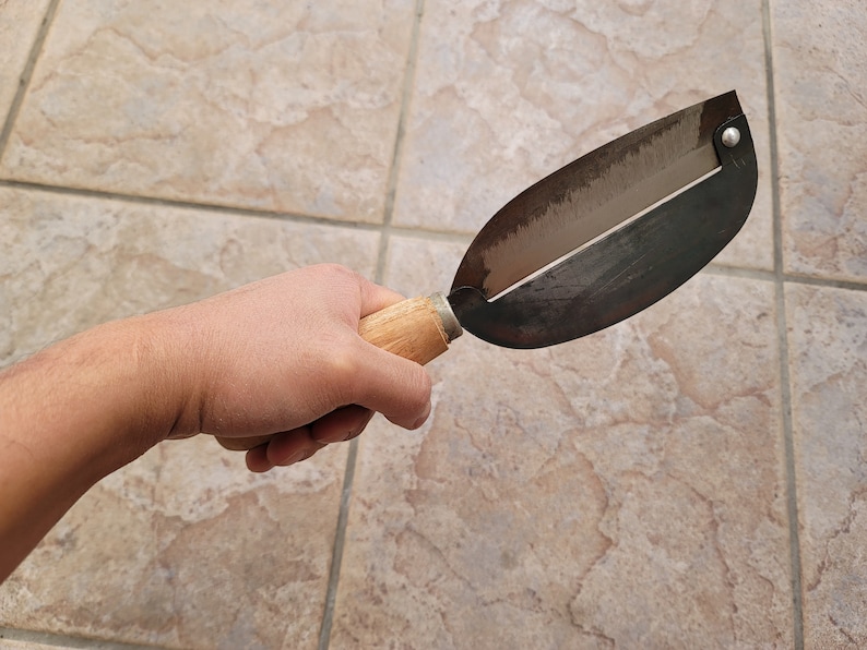 May include: A hand holds a wooden handled metal vegetable peeler with a silver blade. The peeler is on a tiled surface.