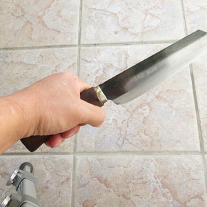 May include: A close-up of a person holding a long, silver knife with a wooden handle. The knife has a slightly curved blade and a dull finish.