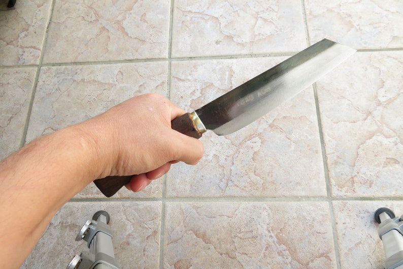 May include: A close-up of a person holding a long, silver knife with a wooden handle. The knife has a slightly curved blade and a dull finish.
