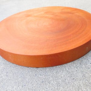 May include: A large, round, wooden cutting board with a reddish-brown hue. The board has a thick profile and visible wood grain patterns, suggesting it is made from a solid piece of wood. The circular shape is ideal for food preparation.