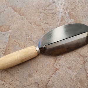 May include: A wooden handled metal vegetable peeler with a silver blade. The peeler is lying on a light brown tiled surface.