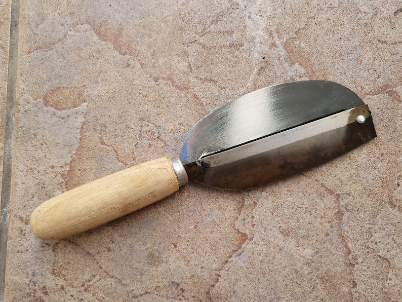 May include: A wooden handled metal vegetable peeler with a silver blade. The peeler is lying on a light brown tiled surface.