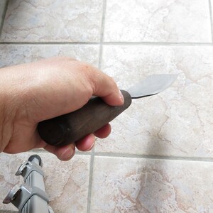 May include: A hand holding a small, wooden-handled, metal scraper tool.