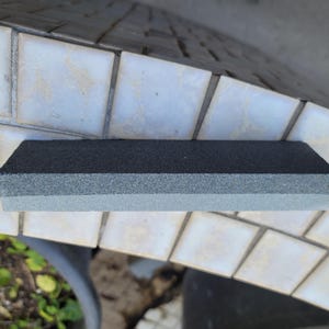 Puede incluir: Una piedra de afilar rectangular con una parte superior gris oscuro y una parte inferior gris claro. La piedra est&aacute; dise&ntilde;ada para afilar cuchillos y herramientas. La imagen muestra la piedra sobre una superficie embaldosada.