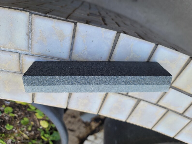 Puede incluir: Una piedra de afilar rectangular con una parte superior gris oscuro y una parte inferior gris claro. La piedra est&aacute; dise&ntilde;ada para afilar cuchillos y herramientas. La imagen muestra la piedra sobre una superficie embaldosada.