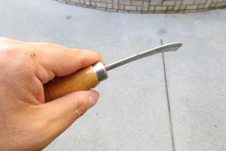 May include: A small, curved blade knife with a wooden handle and a silver metal ferrule. The blade is made of shiny metal. The knife is held in a hand.