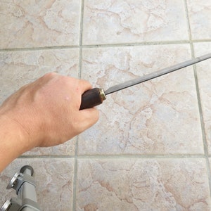 May include: A person holding a long, thin, metal file with a wooden handle. The file has a brass ferrule at the end of the handle.