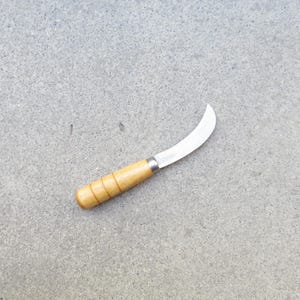 May include: A small, curved blade knife with a wooden handle. The blade is silver and the handle is light brown with a cylindrical shape. The knife is designed for gardening or harvesting.