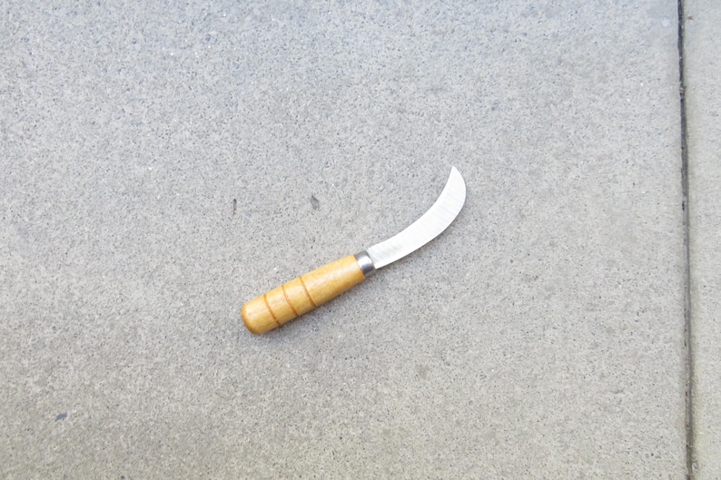 May include: A small, curved blade knife with a wooden handle. The blade is silver and the handle is light brown with a cylindrical shape. The knife is designed for gardening or harvesting.