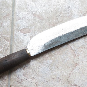May include: A long, silver-colored knife with a wooden handle. The blade is slightly curved and has a dull edge. The handle is dark brown and has a slight grain.