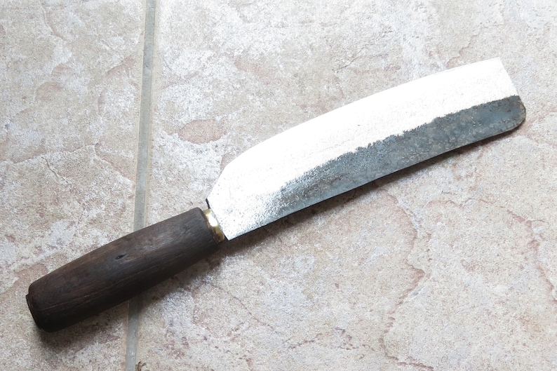 May include: A long, silver-colored knife with a wooden handle. The blade is slightly curved and has a dull edge. The handle is dark brown and has a slight grain.