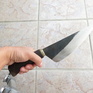 Puede incluir: Una mano sosteniendo un cuchillo grande con mango negro y hoja curva. La hoja est&aacute; parcialmente pulida, mostrando un borde plateado brillante.