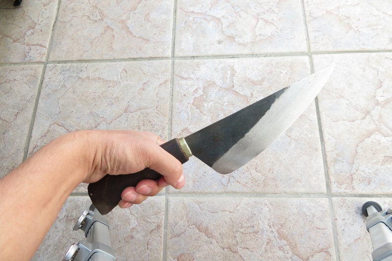 Puede incluir: Una mano sosteniendo un cuchillo grande con mango negro y hoja curva. La hoja est&aacute; parcialmente pulida, mostrando un borde plateado brillante.