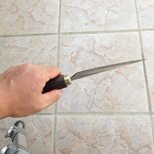 May include: A person holds a long, thin, silver knife with a dark wood handle. The knife has a brass accent near the handle.
