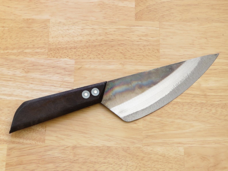 May include: A black handled knife with a silver blade and two rivets on the handle. The knife is lying on a wooden surface.