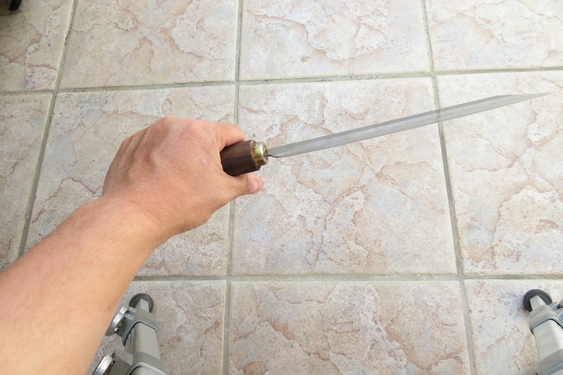 Peut inclure: Une personne tient une lime m&eacute;tallique longue et fine avec une poign&eacute;e en bois. La lime est tenue horizontalement avec la poign&eacute;e sur le c&ocirc;t&eacute; droit de l'image. La lime est tranchante et pr&eacute;sente une l&eacute;g&egrave;re courbure.