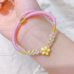 May include: A pink and white string bracelet with a gold flower charm. The bracelet is made of braided string and has a knot closure.