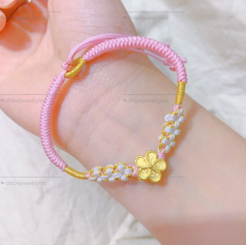 May include: A pink and white string bracelet with a gold flower charm. The bracelet is made of braided string and has a knot closure.