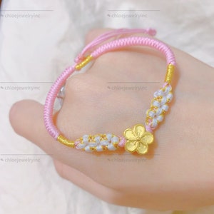 May include: A pink and gold braided bracelet with a gold flower charm. The bracelet is made with a knotting technique and has a white flower accent.