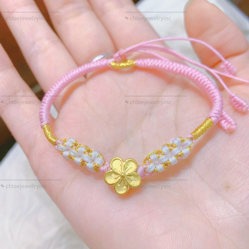 May include: A pink and gold braided bracelet with a gold flower charm. The bracelet is made with a knotting technique and has white flower accents.