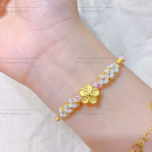 May include: A pink and white string bracelet with a gold flower charm. The bracelet is tied in a knot pattern.