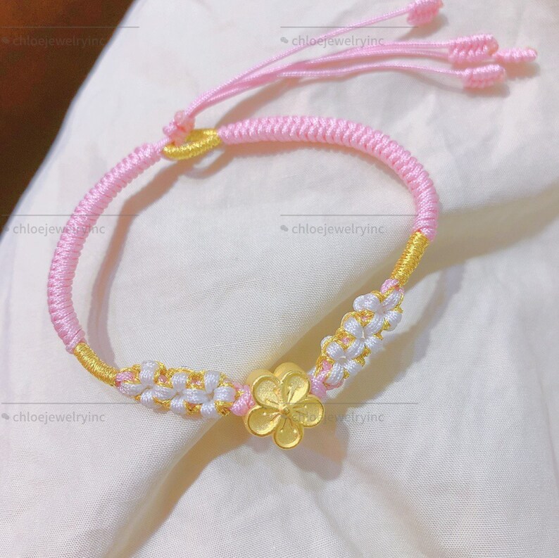 May include: A pink and gold braided bracelet with a gold flower charm. The bracelet is made of string and has a knot at the end.
