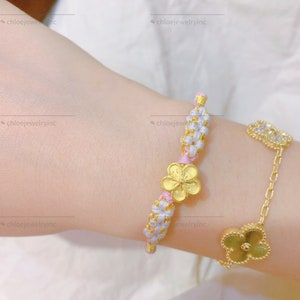 May include: A pink and white braided bracelet with a gold flower charm. The bracelet is on a person's wrist.
