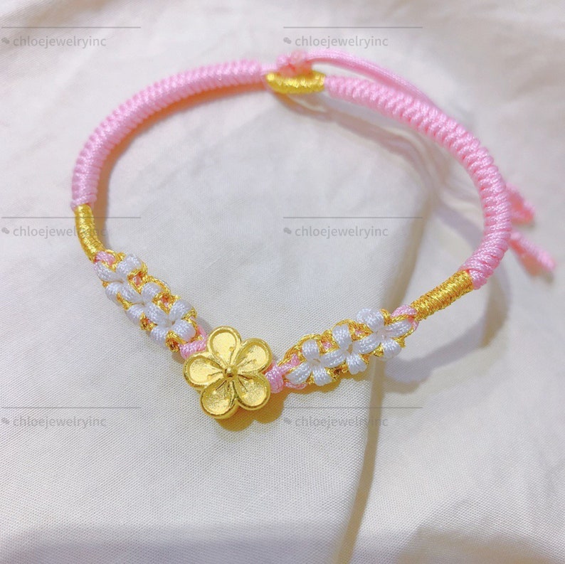 May include: A pink and white braided string bracelet with a gold flower charm. The bracelet is tied with a knot and has a tassel on the end.