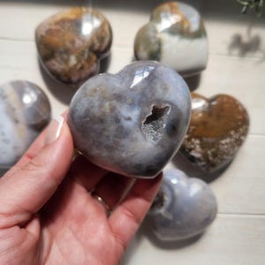 May include: Heart-shaped polished stones in shades of gray, brown, and white. One stone is held in a hand, revealing a cavity with crystal formations. The other stones are scattered around the hand. The stones have a natural, organic appearance.