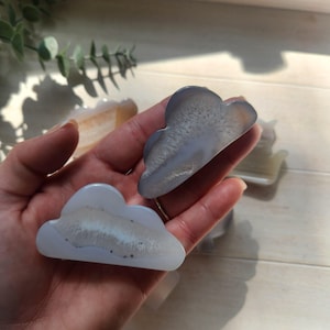 May include: Cloud-shaped agate stones in shades of gray and white. The polished stones have a natural, layered appearance, with a cloudy pattern. The stones are held in a hand, with other similar stones visible in the background. These stones are ideal for home decor.