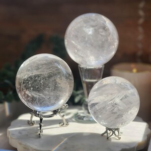 May include: Three clear quartz crystal spheres of varying sizes. The largest sphere rests on a tall, clear glass pedestal, while the two smaller spheres are supported by intricate, silver-toned metal stands with ornate, curved legs.