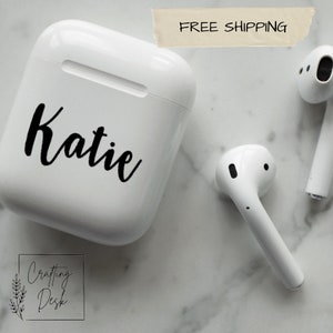 May include: White Airpods case with a black vinyl sticker that says "Katie" on it. The case is on a white marble surface.
