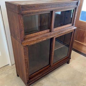 May include: A dark brown wooden bookcase with two sections, each with sliding glass doors. The bookcase has a rectangular shape and is made of wood with visible grain. The top section is smaller than the bottom section.