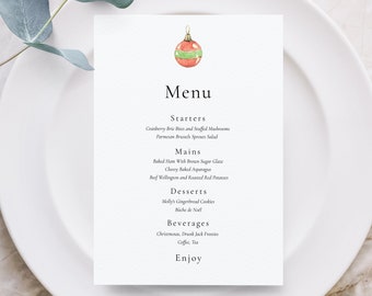 May include: A white menu card with a watercolor Christmas ornament illustration at the top, placed on a white plate. The menu lists starters, mains, desserts, and beverages. The menu is for a Christmas dinner.