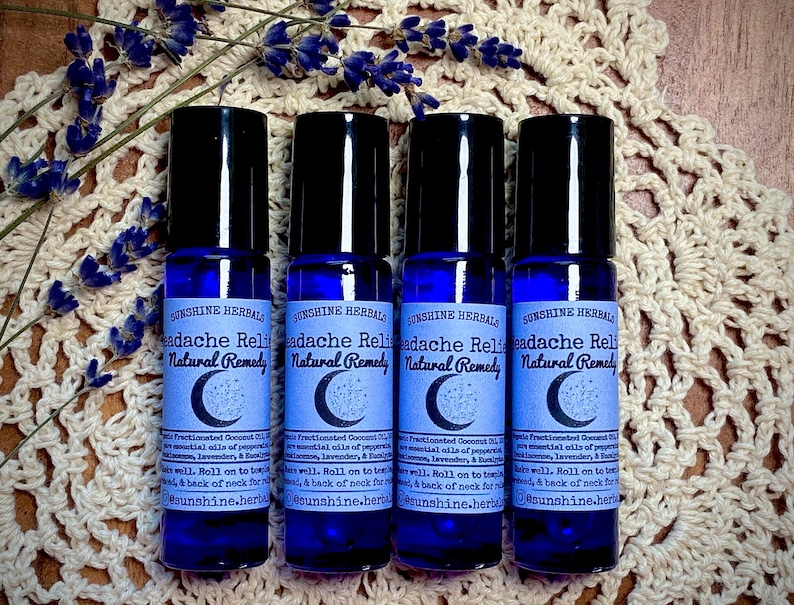 Puede incluir: Cuatro botellas de vidrio azul con rodillo y una etiqueta blanca que dice "Sunshine Herbals Headache Relief Natural Remedy". Las botellas est&aacute;n dispuestas en fila sobre una superficie de crochet blanco.