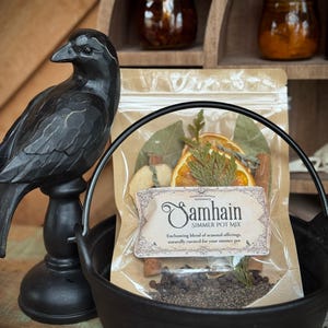 May include: A black cast iron cauldron holds a bag of Samhain Simmer Pot Mix, with dried orange slices, cinnamon sticks, and other botanicals. A black raven statue stands to the left. Two jars of preserves sit on a wooden shelf in the background.
