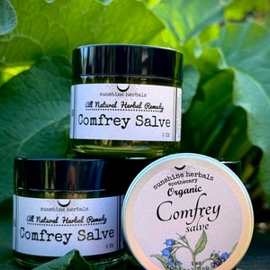 May include: Two jars of Comfrey Salve, one labeled "All Natural Herbal Remedy" and the other labeled "Organic Comfrey Salve". Both jars have a black lid and a green label with a white crescent moon and the words "sunshine herbals" printed on them.