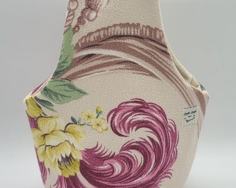 May include: A beige fabric tote bag with a floral print featuring pink, yellow, and green flowers. The bag has a small white label with a simple face design.